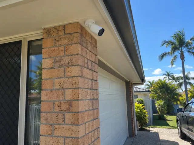 White CCTV camera mounted under a house eave beside a brick garage wall in Mermaid Waters